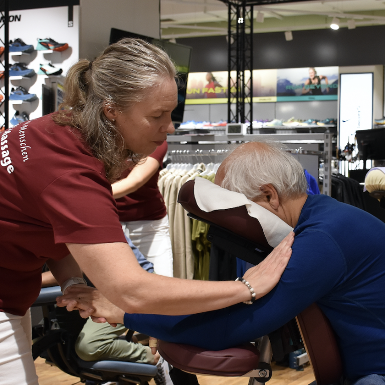 TouchLife® Mobile Massage Eine Frau hilft einem älteren Mann, der auf einem Stuhl sitzt, mit einer mobilen Massage